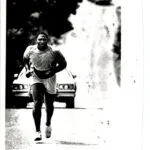 Runner jogging on road with car and building in background, black and white photograph