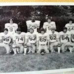 Black and white photo of high school football team with eleven players wearing Warriors uniforms with numbers, posed on grass with trees in background