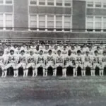 Black and white photograph of a football team posing in rows on a field, wearing numbered uniforms in front of a brick building.