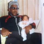 Man wearing sunglasses and headband holding an infant in white clothing indoors