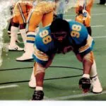 Football player wearing blue and orange uniform with number 98 in defensive stance on field