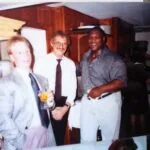 Three men standing together indoors at what appears to be a social gathering, with wood paneling and shelving visible in the background.