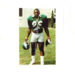 Football player in green and white uniform holding helmet on grass field with building in background