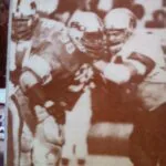 Sepia-toned vintage photograph of football players in helmets and uniforms gathered on field