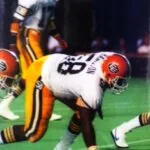 Football player in white and orange uniform bent in defensive stance on field during game