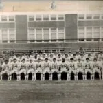 Black and white team photo of football players in numbered uniforms seated in rows with stadium and brick building in background