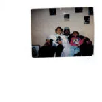 Three people sitting together on a couch indoors, with photographs visible on the wall behind them.