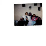 Three people sitting together on a couch in an indoor room with photos on the wall behind them.